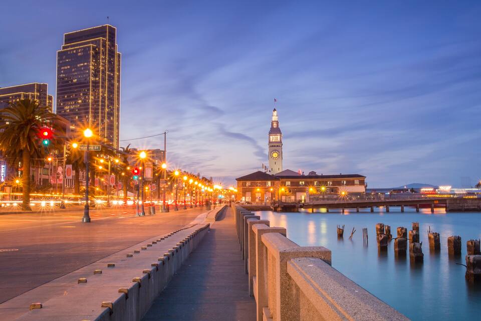 Una passejada al costat del riu amb una torre del rellotge, edificis il·luminats i un riu tranquil.