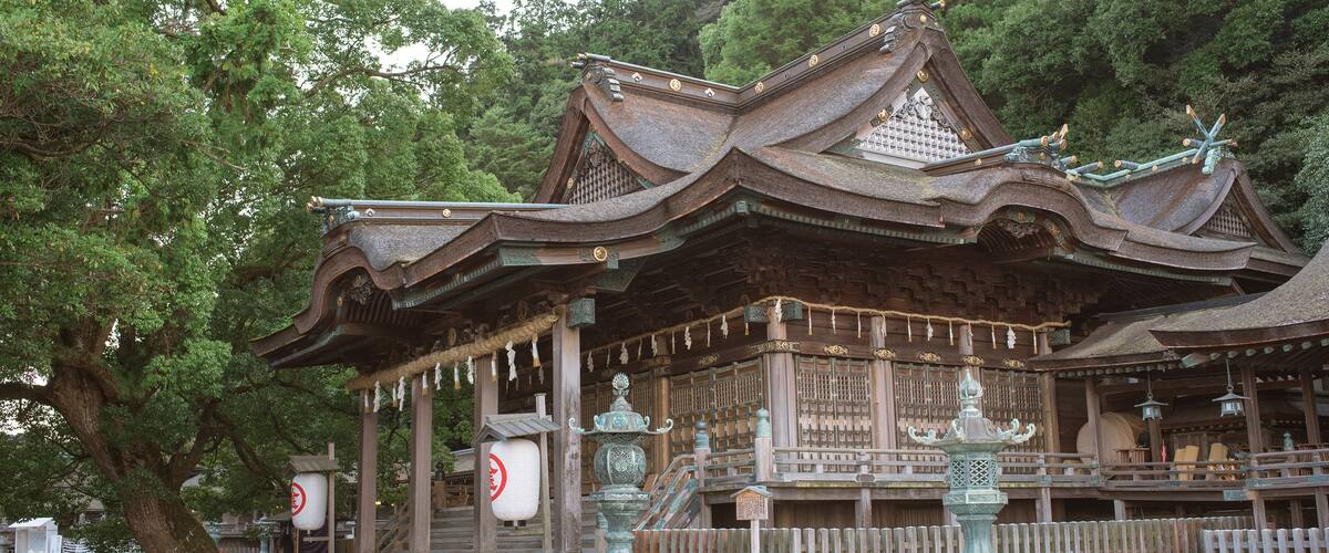 A traditional Japanese temple with ornate wooden architecture, a large tree to the left, and a wooden fence in the foreground.