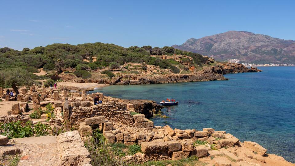 Ancient ruins by a coastal area with clear blue water and mountains in the background.