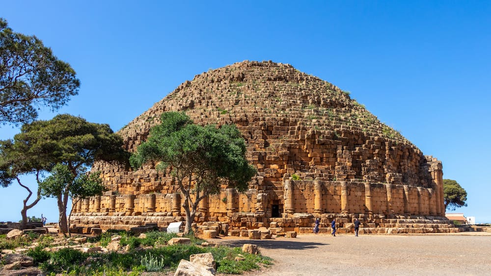 An ancient stone structure with columns and a dome, surrounded by trees and rocky terrain.