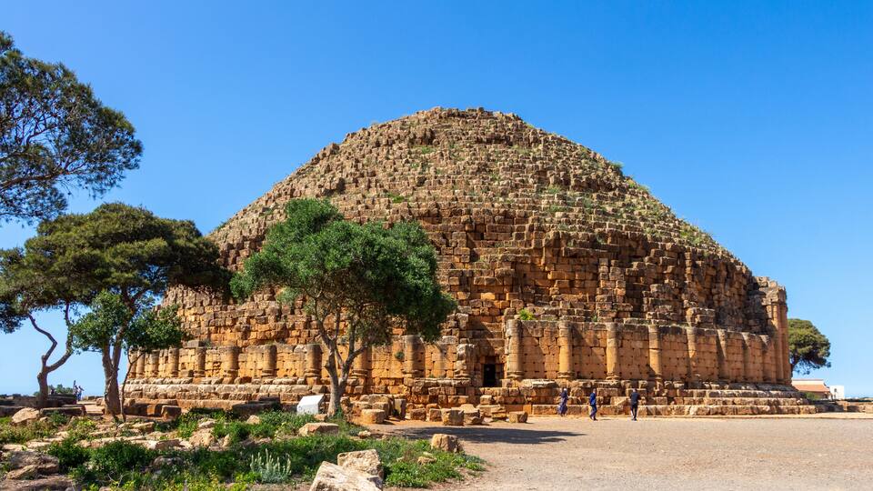 An ancient stone structure with columns and a dome, surrounded by trees and rocky terrain.