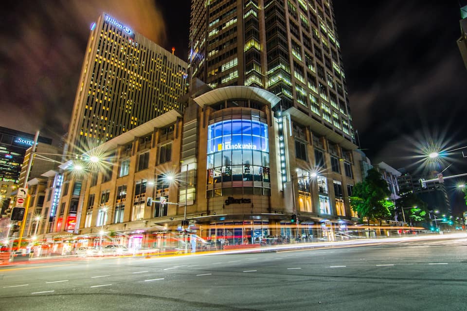 A nighttime cityscape with illuminated skyscrapers, a prominent glass-fronted building, and a busy street with light trails from moving vehicles.