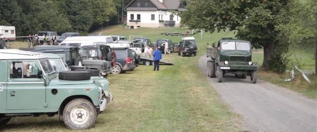 A rural road with parked vehicles, a house in the background, and a grassy area.