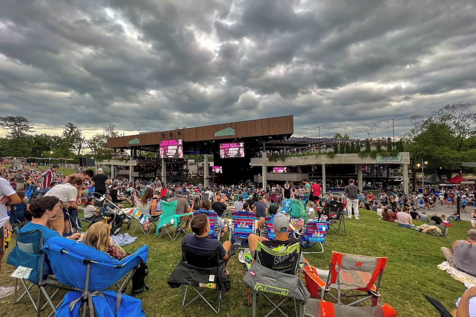 A crowded outdoor event with a stage and large screens, surrounded by grassy areas and trees.