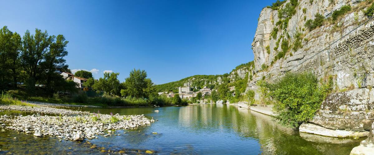 A river with clear water, surrounded by rocky banks and greenery, with a village on a hillside in the background.