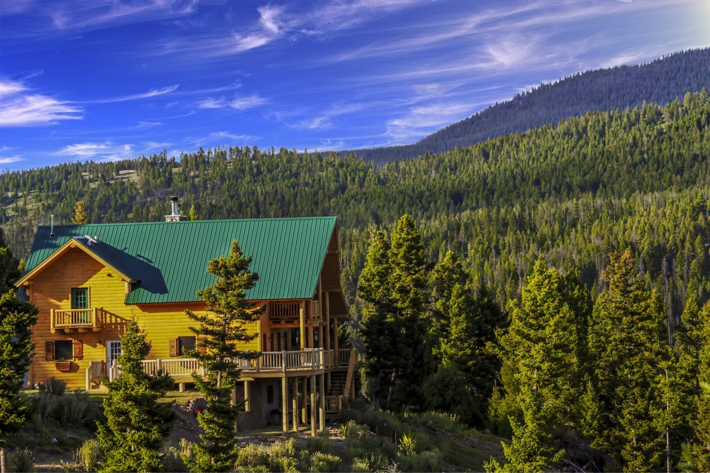 A wooden cabin with a green roof surrounded by dense forest and mountains.