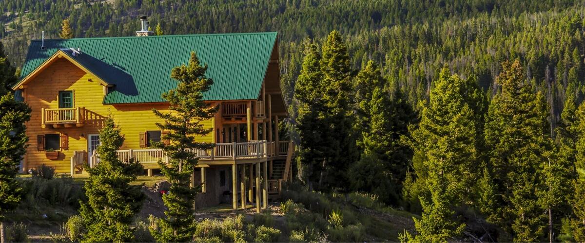 A wooden cabin with a green roof surrounded by dense forest and mountains.
