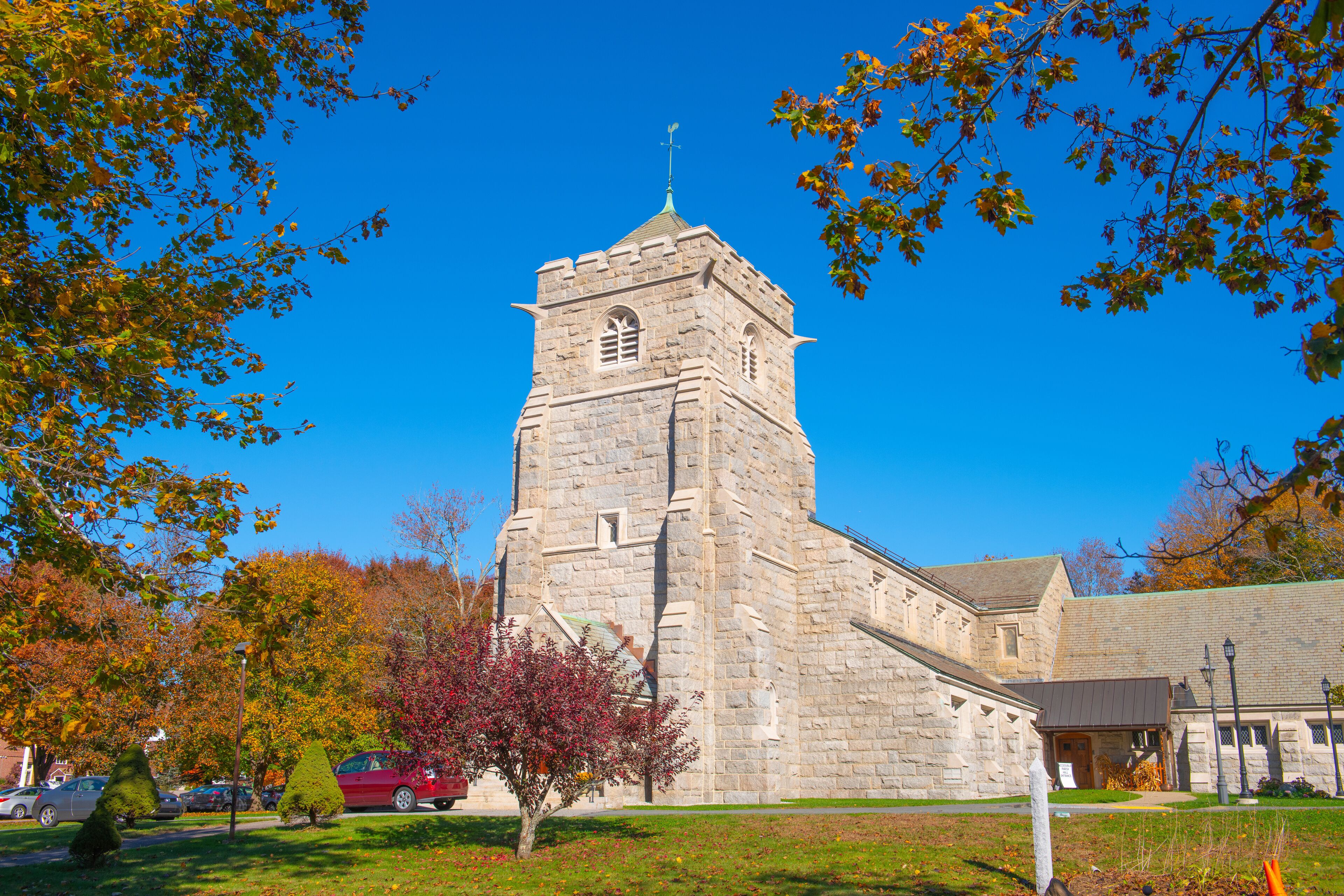 A stone church with a bell tower, surrounded by autumn-colored trees and a clear blue sky.