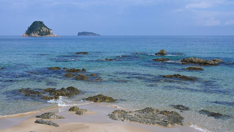 A beach with clear turquoise water, rocky outcrops, and distant islands.