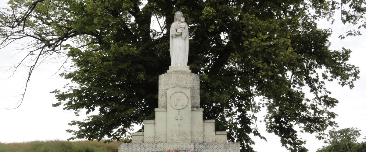 A stone monument with a cross and a statue of a woman holding a child, surrounded by greenery.