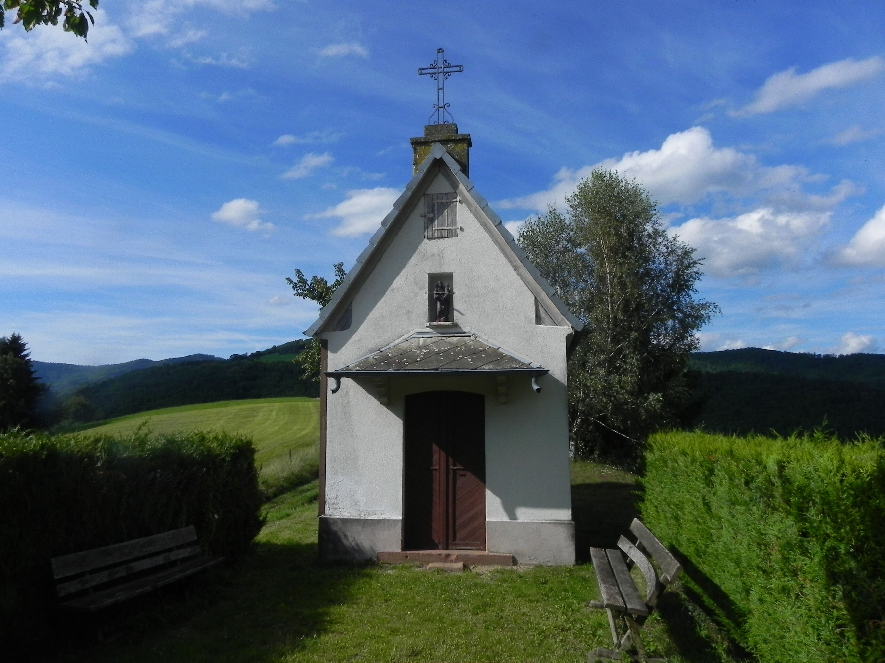 A small white church with a cross on top, surrounded by green fields and hills.