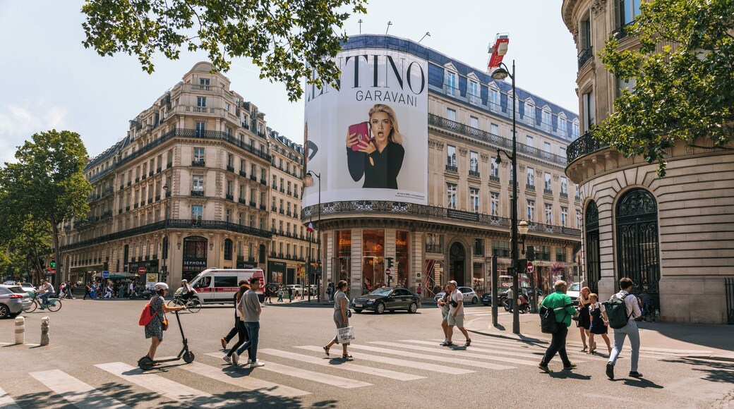 En travl bygningvej med fodgængere, køretøjer og et stort TINO GARAVANI reklameskilt.