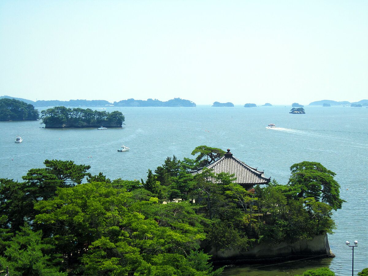 A traditional Japanese temple surrounded by lush greenery and a calm sea with boats.