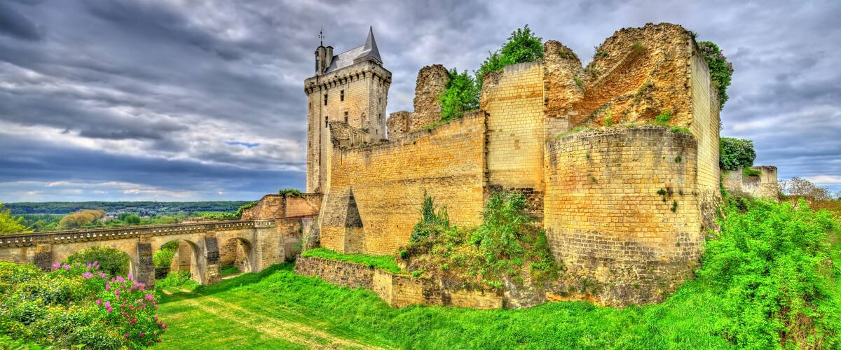 A historic castle with a large tower, surrounded by lush greenery and a stone bridge.