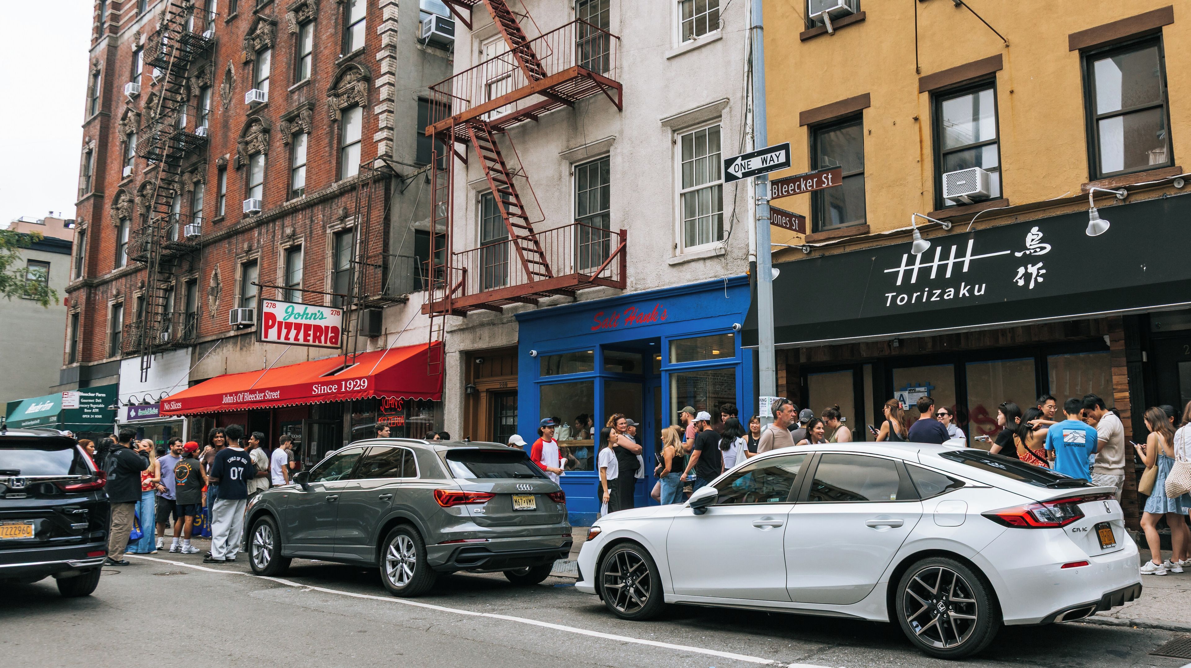 A busy street with cars, pedestrians, and various storefronts including a pizzeria and a restaurant.