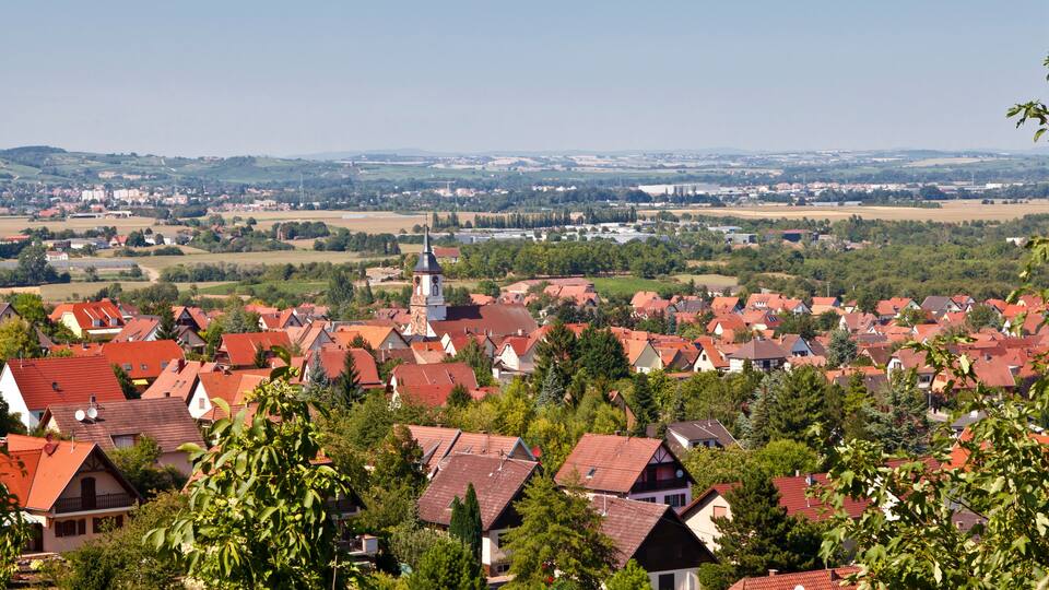 A town with red-roofed houses, a church with a clock tower, and a river in the background.