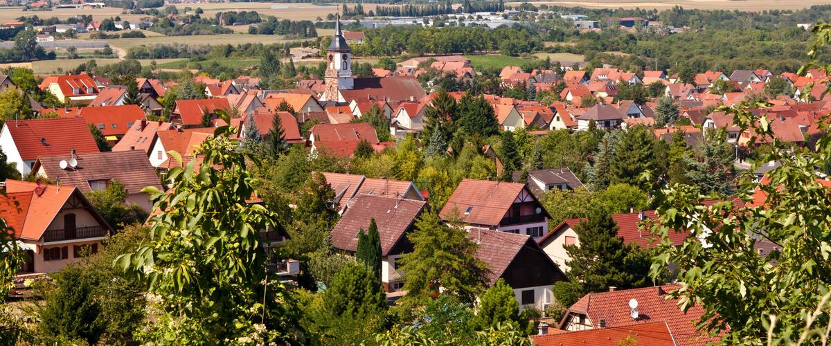 A town with red-roofed houses, a church with a clock tower, and a river in the background.