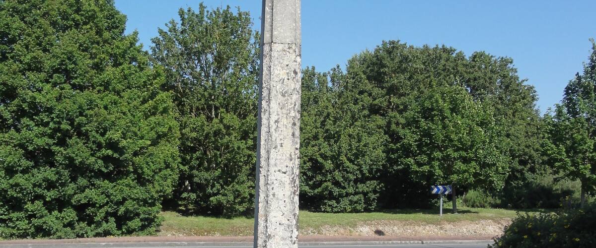 A weathered stone cross on a pedestal in a grassy area with trees in the background.