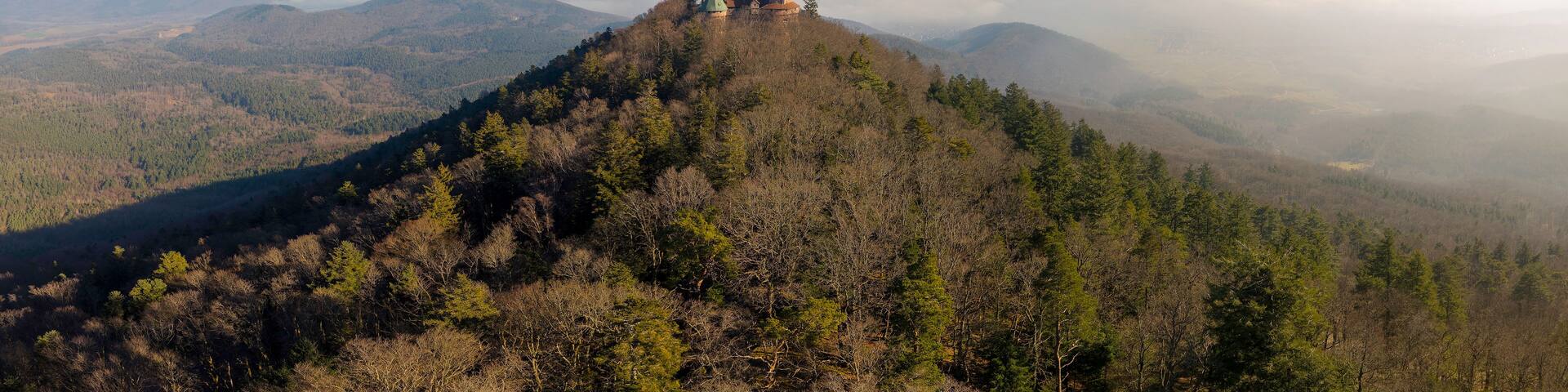 A mountain with a castle on top, surrounded by forested hills and a clear sky.