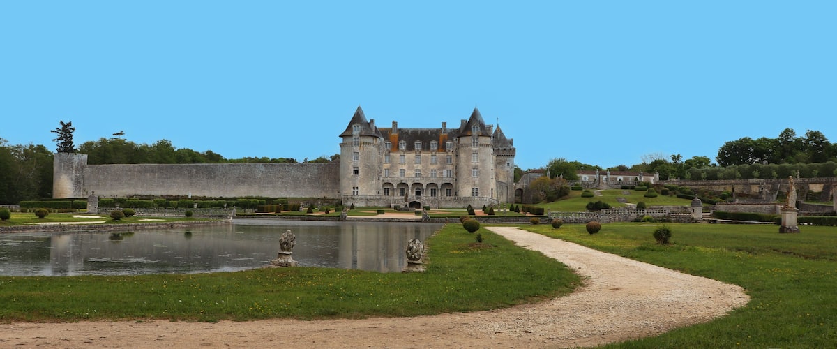 A historic castle with a moat, surrounded by manicured gardens and a clear blue sky.