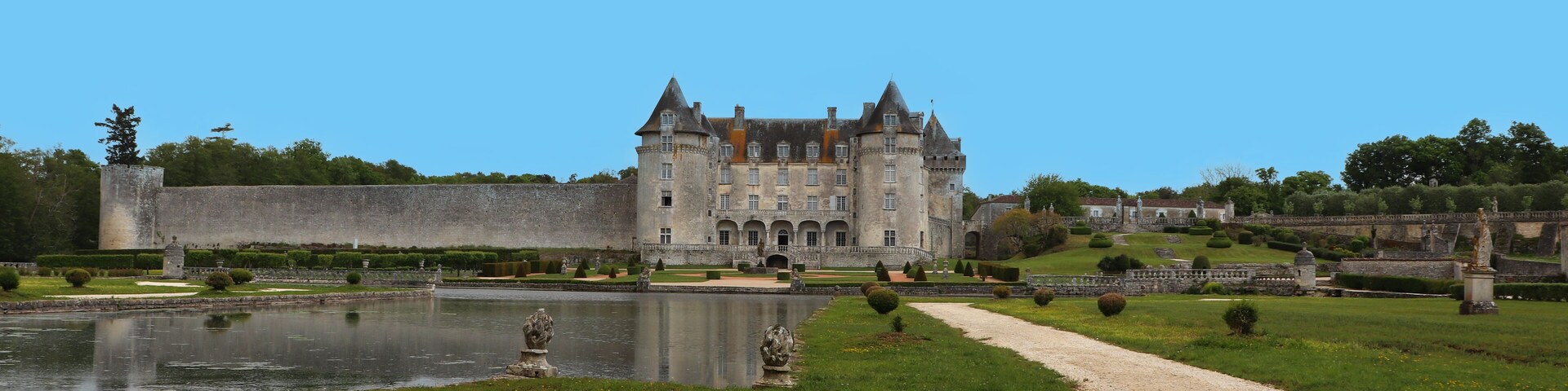 A historic castle with a moat, surrounded by manicured gardens and a clear blue sky.