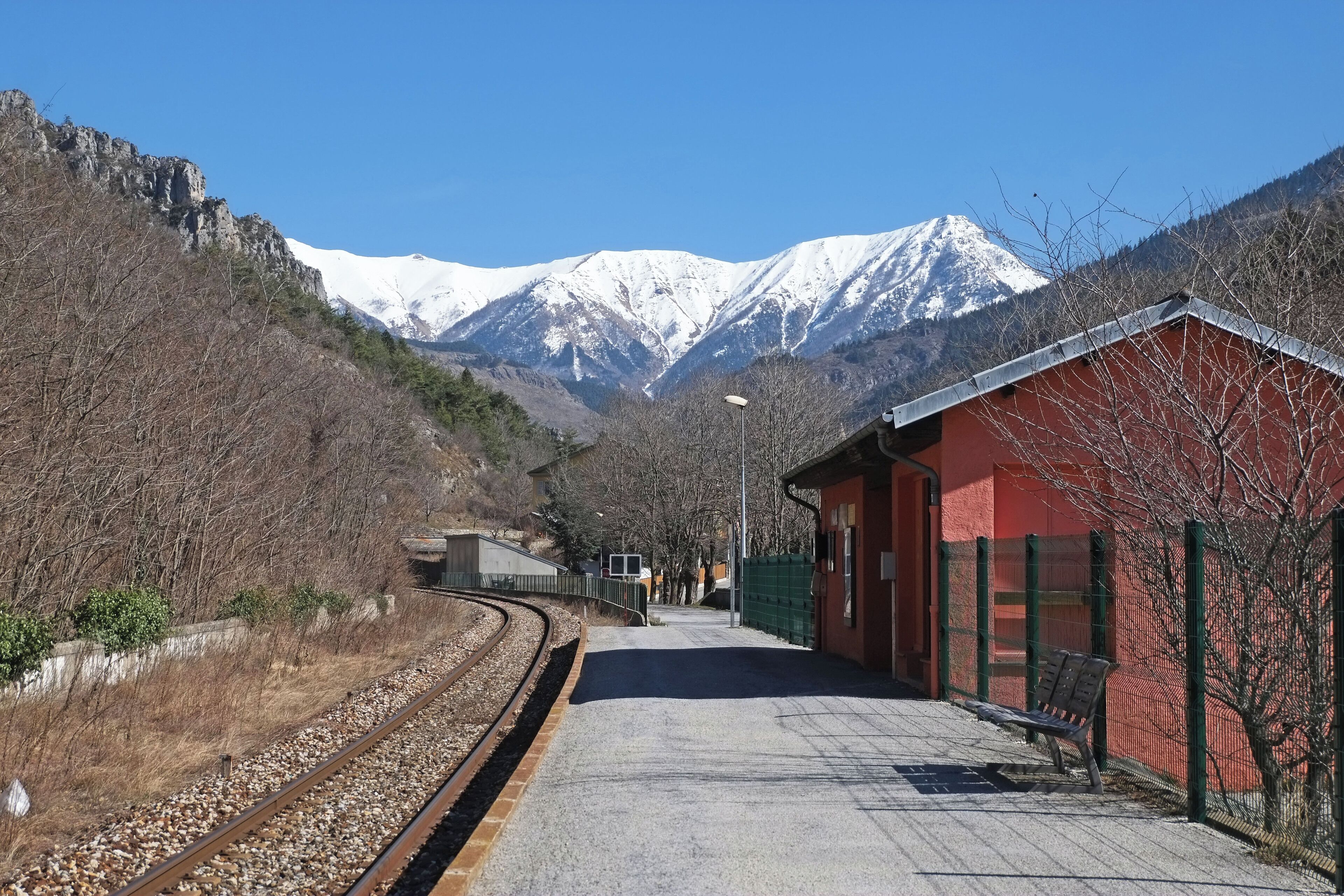 A railway station with a red building, tracks, and snow-capped mountains in the background.