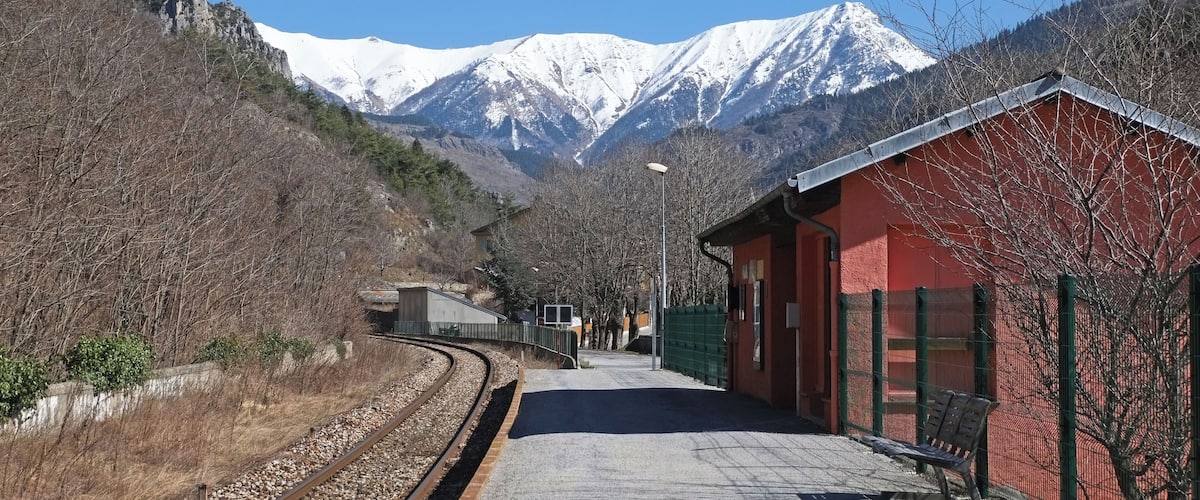 A railway station with a red building, tracks, and snow-capped mountains in the background.