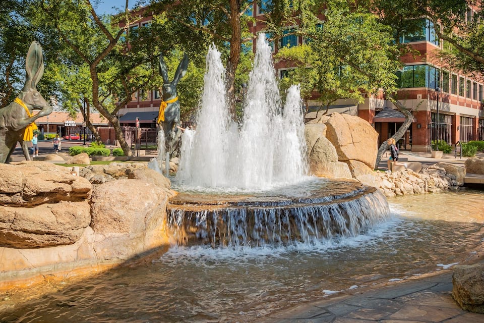 Ein Brunnen mit mehreren Wasserstrahlen, umgeben von Steinen und Bäumen, mit Gebäuden im Hintergrund.