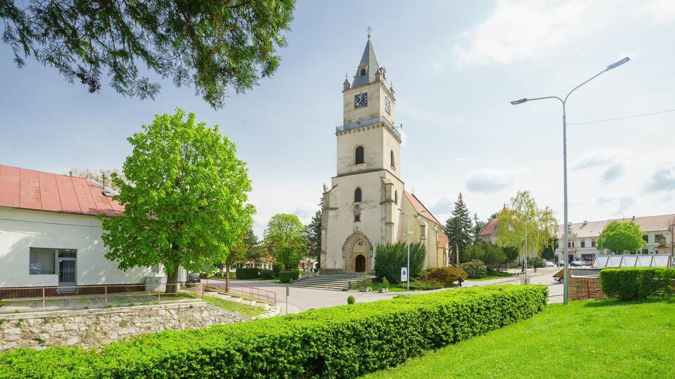 A church with a tall tower and a clock, surrounded by greenery and a paved street.