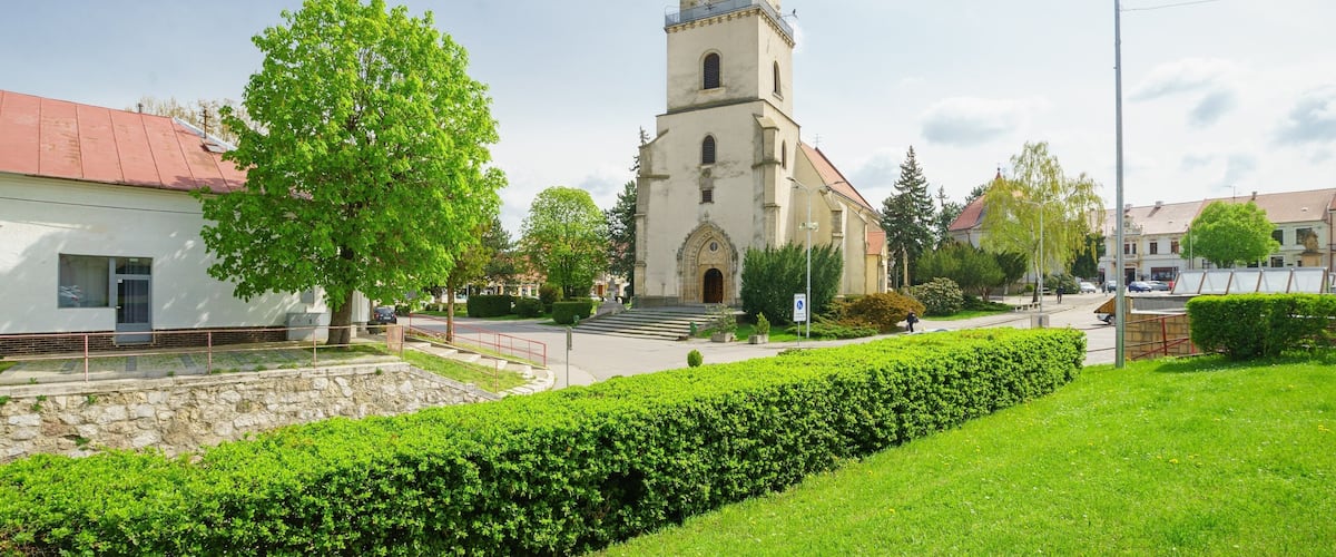 A church with a tall tower and a clock, surrounded by greenery and a paved street.