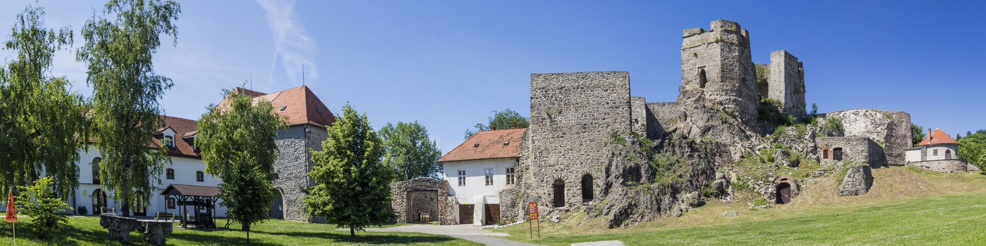 A historic stone castle with a tower, surrounded by lush green grass and a white building with a red roof.