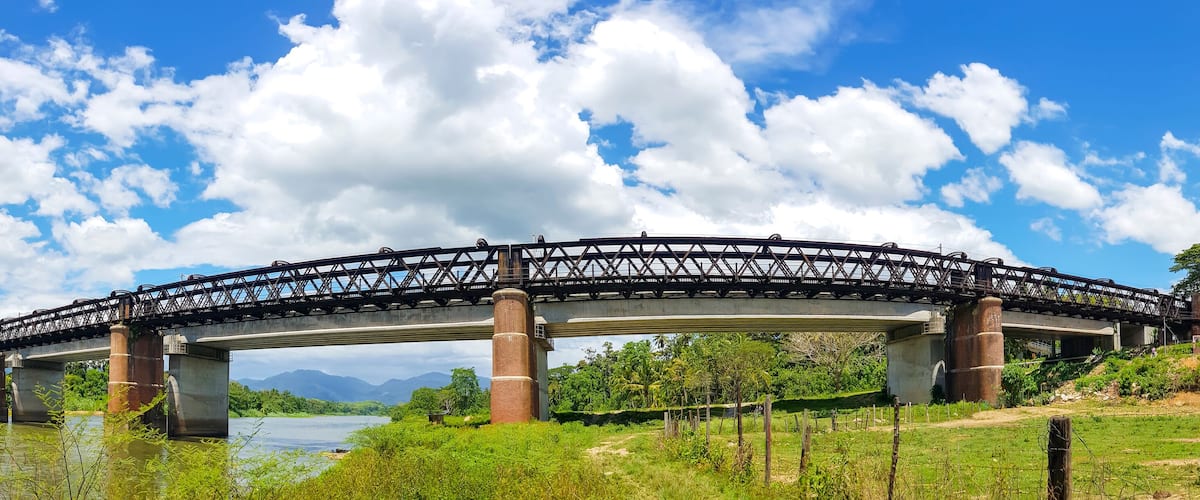 A steel bridge over a river with mountains in the background and a clear sky.