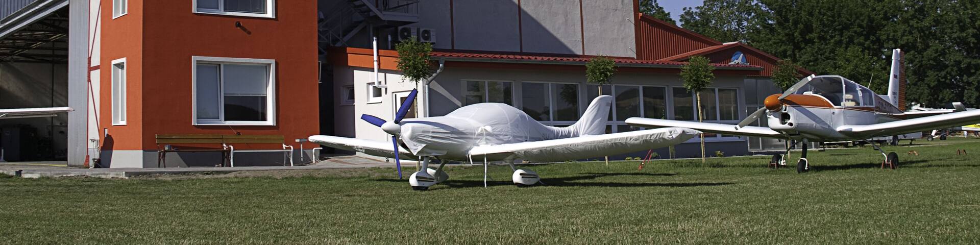 A building with a sign reading "PARTIZANSKE" and several small aircraft parked on the grass in front.