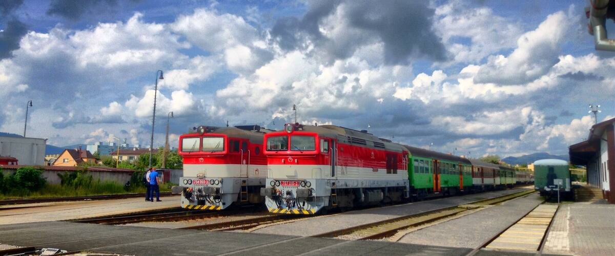 A train station with two trains on the tracks, a clear sky, and some buildings in the background.