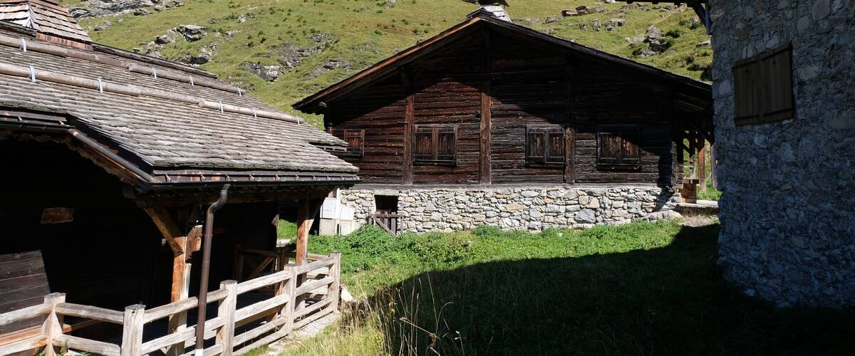 A wooden cabin with a stone wall, surrounded by a wooden fence and a forested hillside.