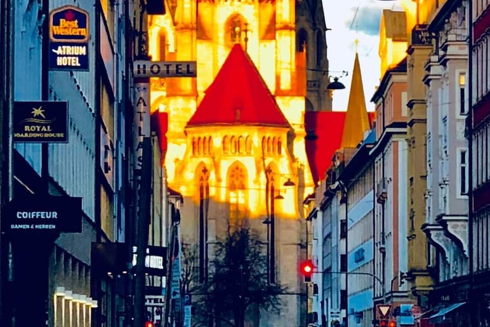 A historic church with a tall spire and illuminated red roof, surrounded by urban buildings and parked cars.