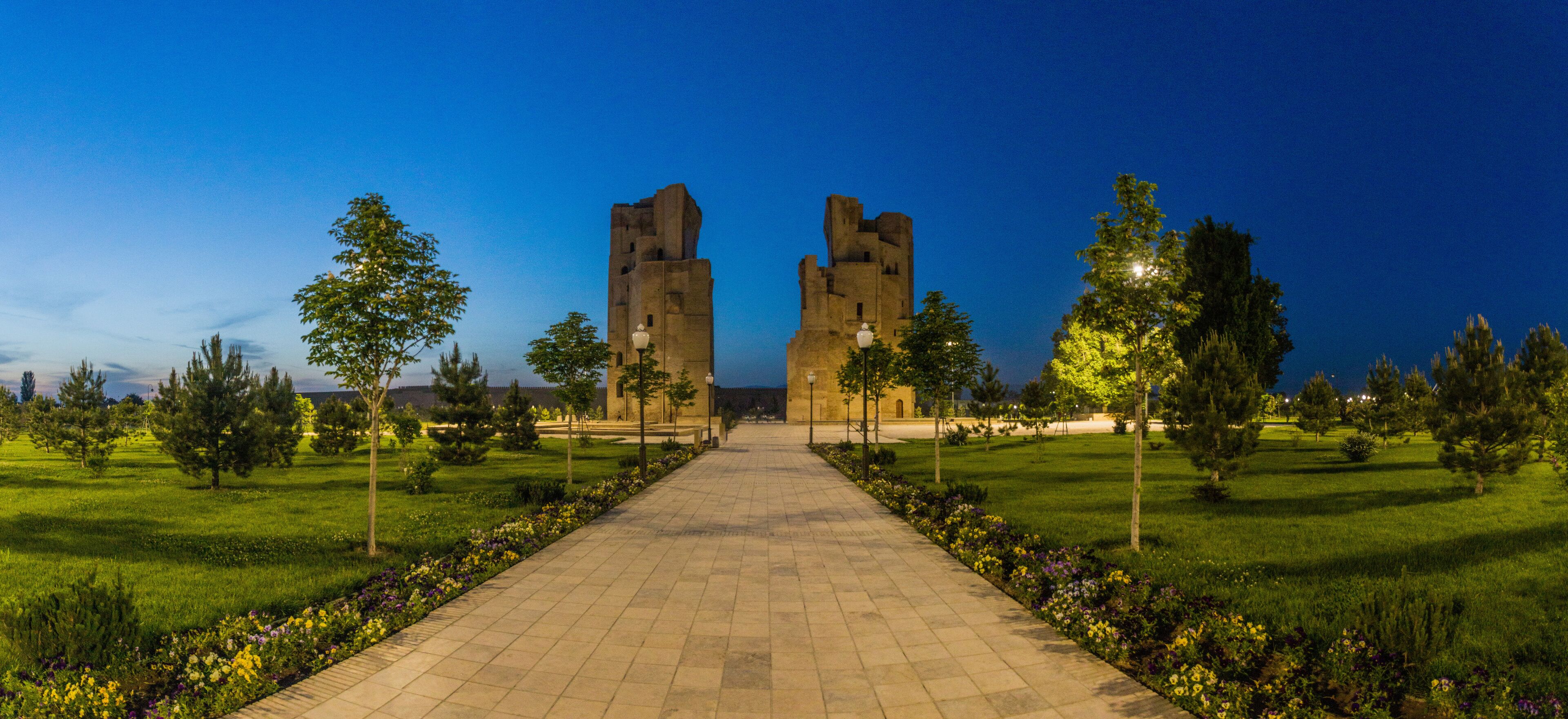 A pathway leading to a castle-like structure with two towers, surrounded by a well-maintained garden and trees.