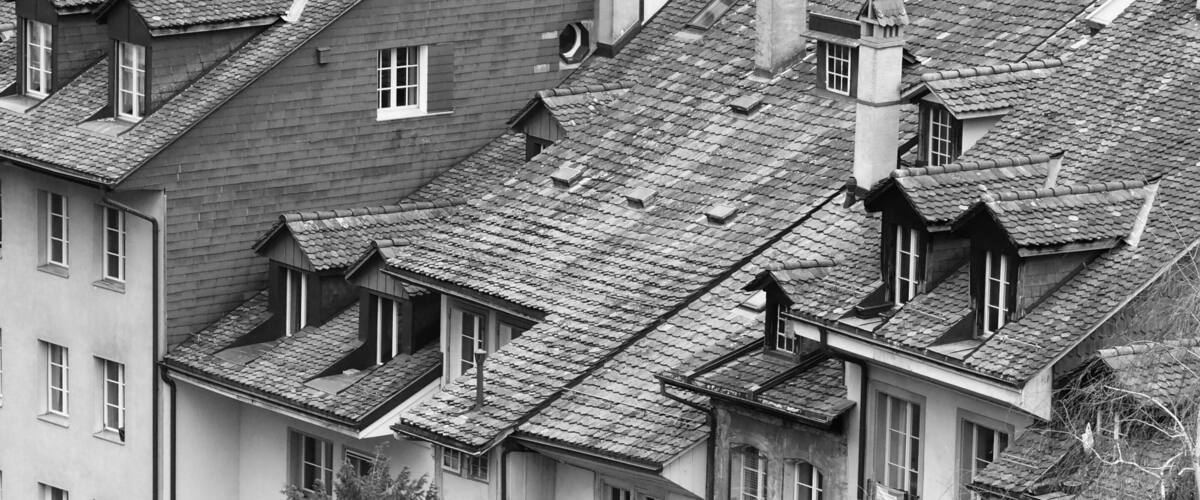 A black and white view of a densely packed residential area with traditional European-style buildings, steeply pitched roofs, and a prominent tree in the foreground.
