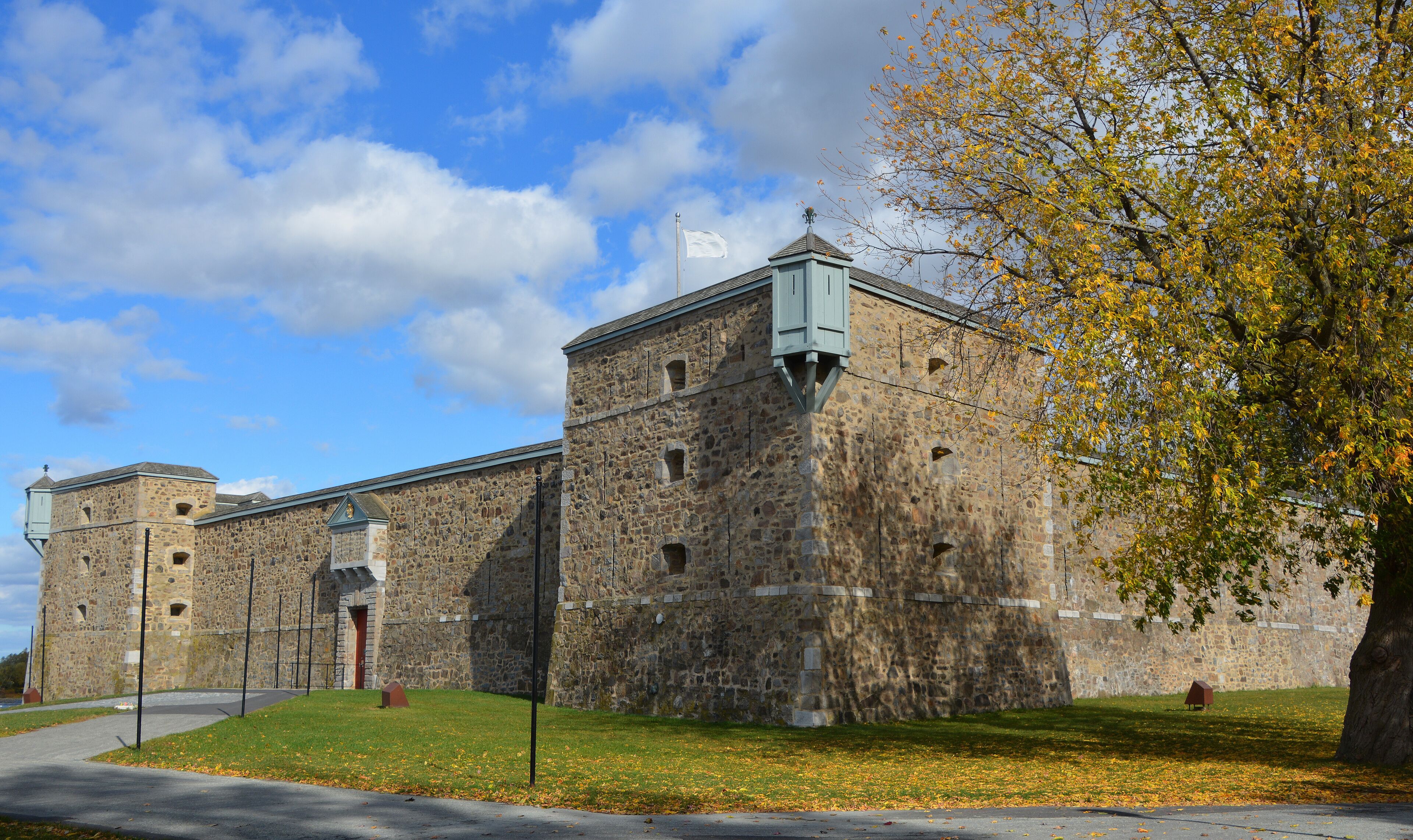 A historic stone building with a flagpole, a flag, and a tree with autumn leaves.