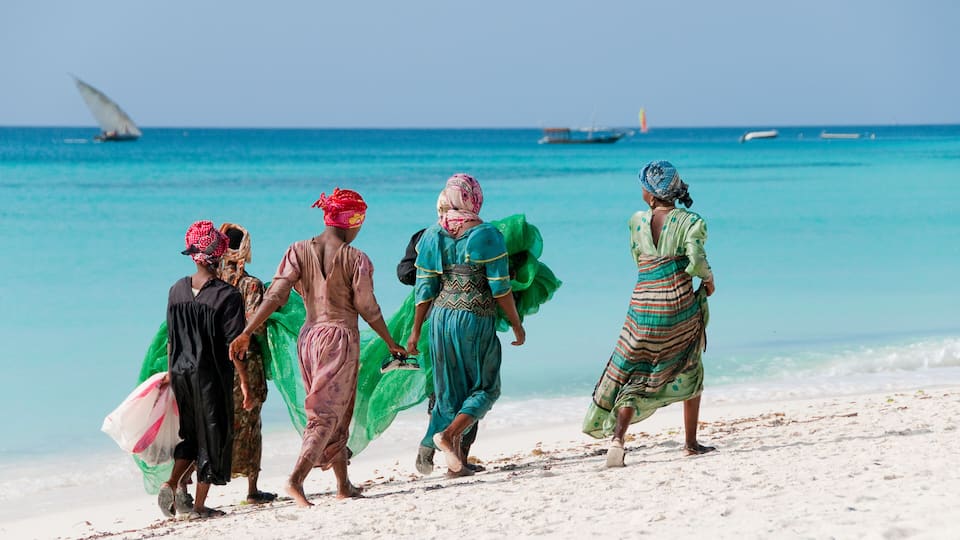 A group of people walking on a sandy beach with the ocean and boats in the background.