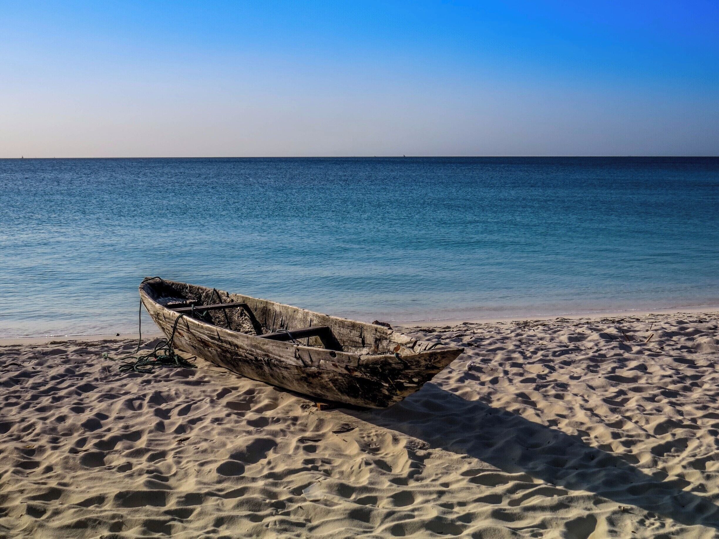 A weathered boat on a sandy beach with calm blue waters in the background.