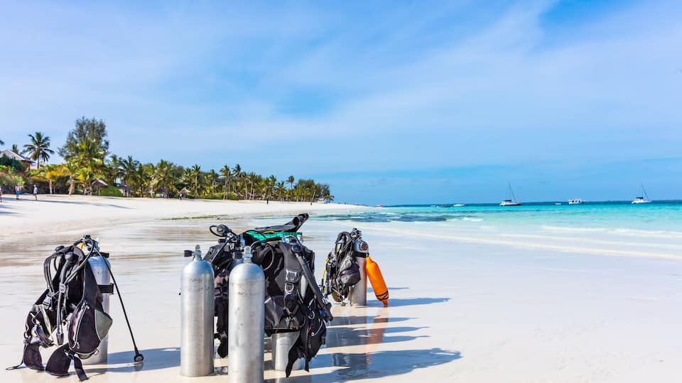 Scuba diving gear on a beach with clear blue water and boats in the distance.