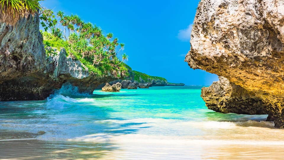 A tropical beach with clear turquoise waters, rocky cliffs, and lush greenery.
