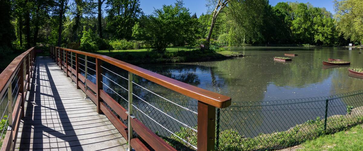 A wooden bridge over a calm river with boats, surrounded by lush greenery and trees.