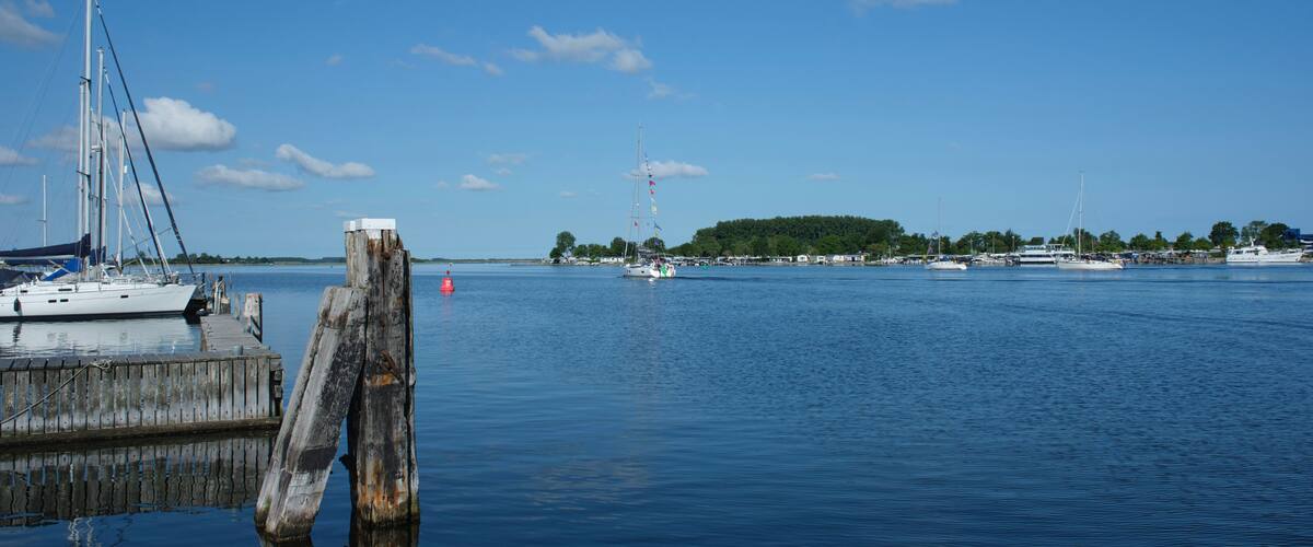 A marina with sailboats and yachts docked, a wooden pier, and a distant island with a lighthouse.
