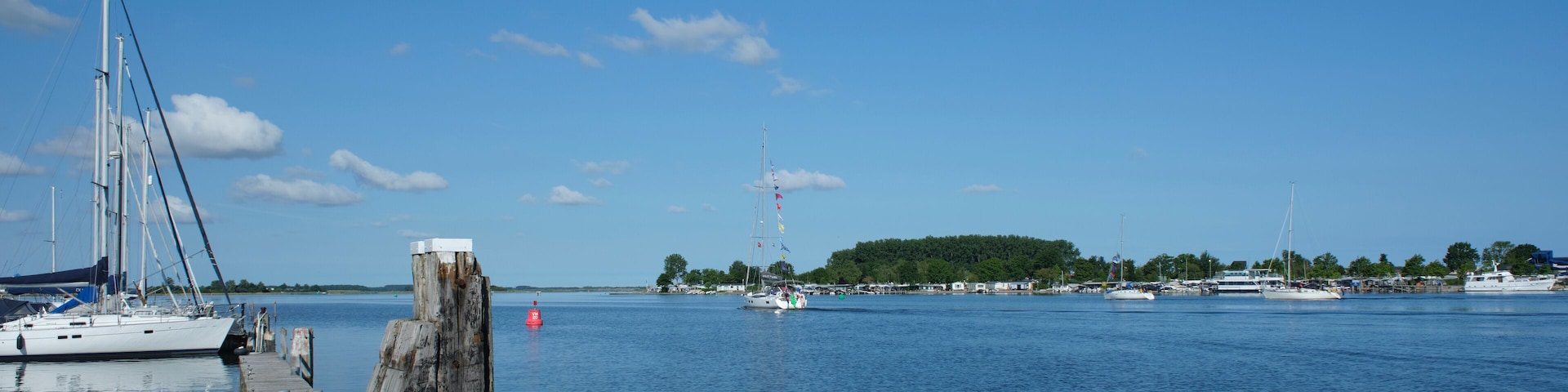 A marina with sailboats and yachts docked, a wooden pier, and a distant island with a lighthouse.