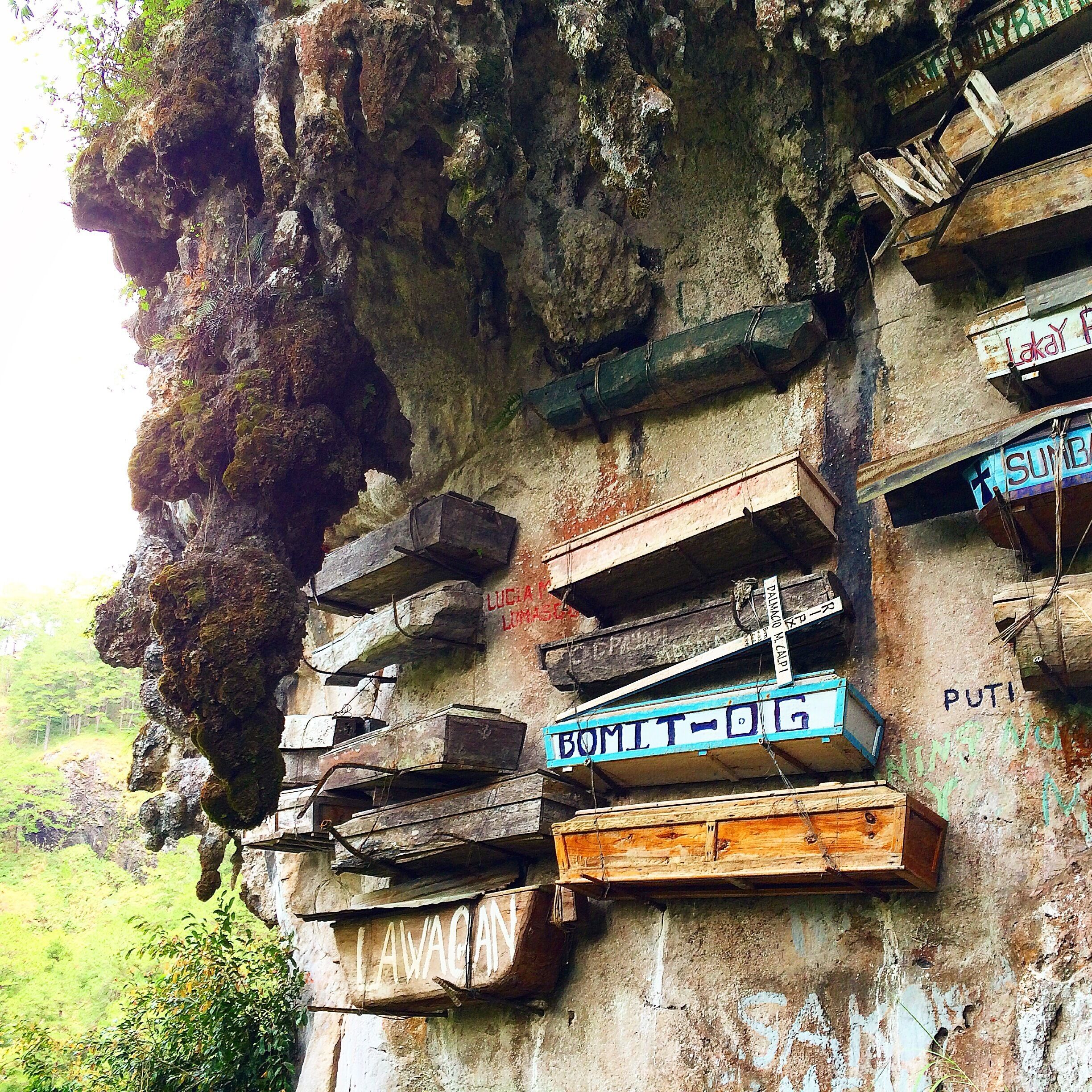 Wooden signs attached to a rocky cliff, with names like 'LUCIA LUCAS' and 'BOMITOG' visible.