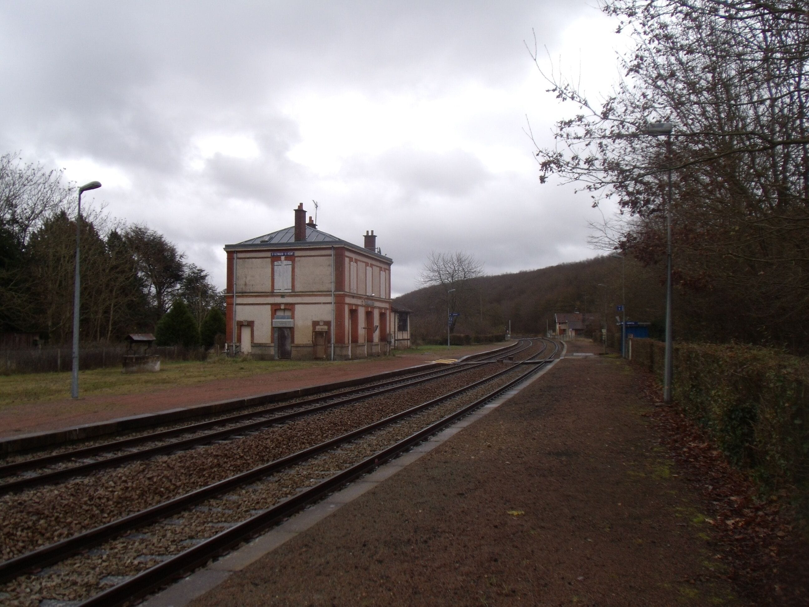 A railway station with tracks, a building, and a hilly landscape in the background.