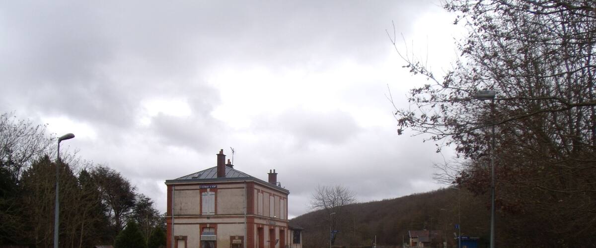 A railway station with tracks, a building, and a hilly landscape in the background.