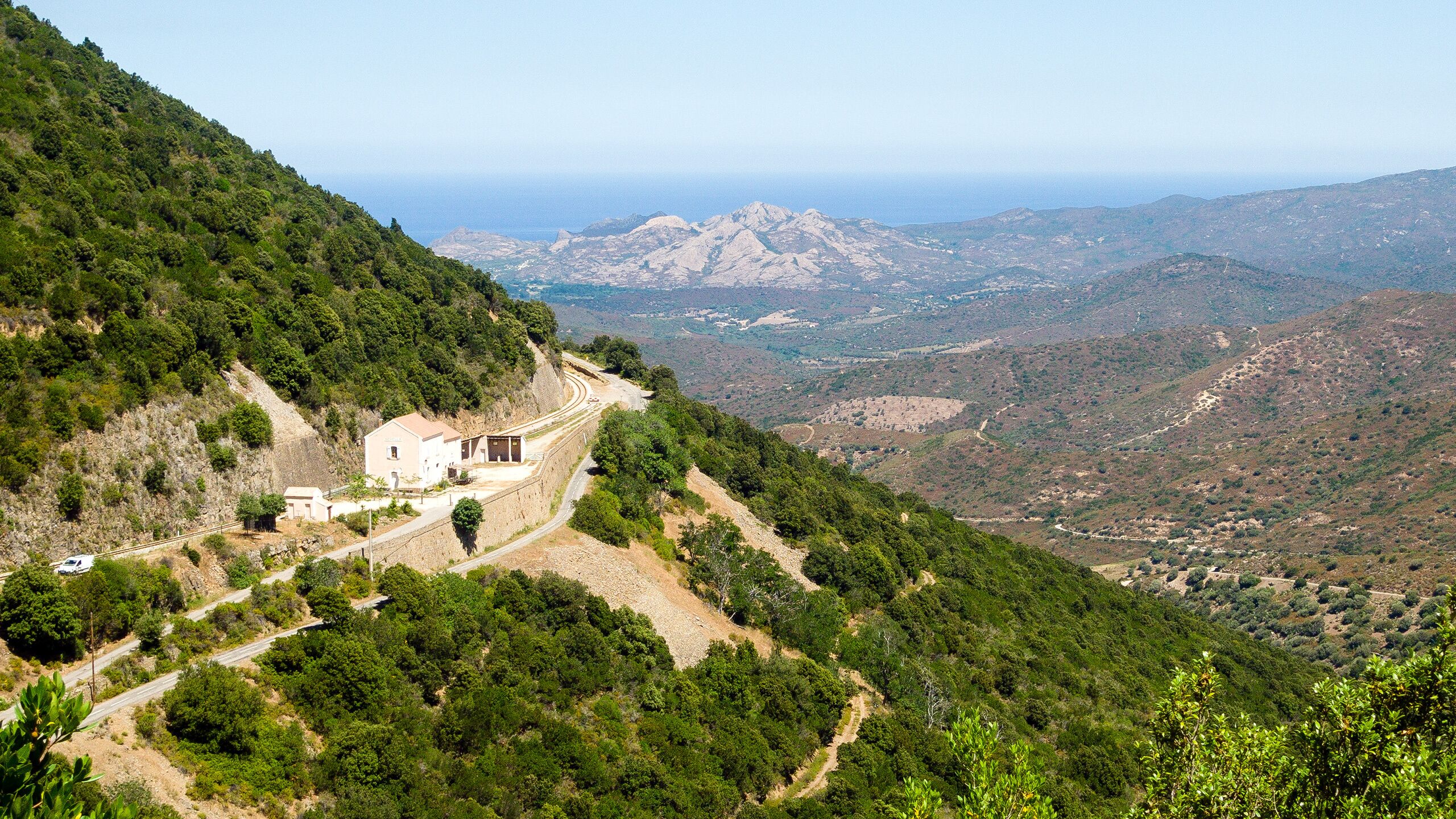 A winding road through a mountainous landscape with a small building on the left and a view of distant mountains and valleys.